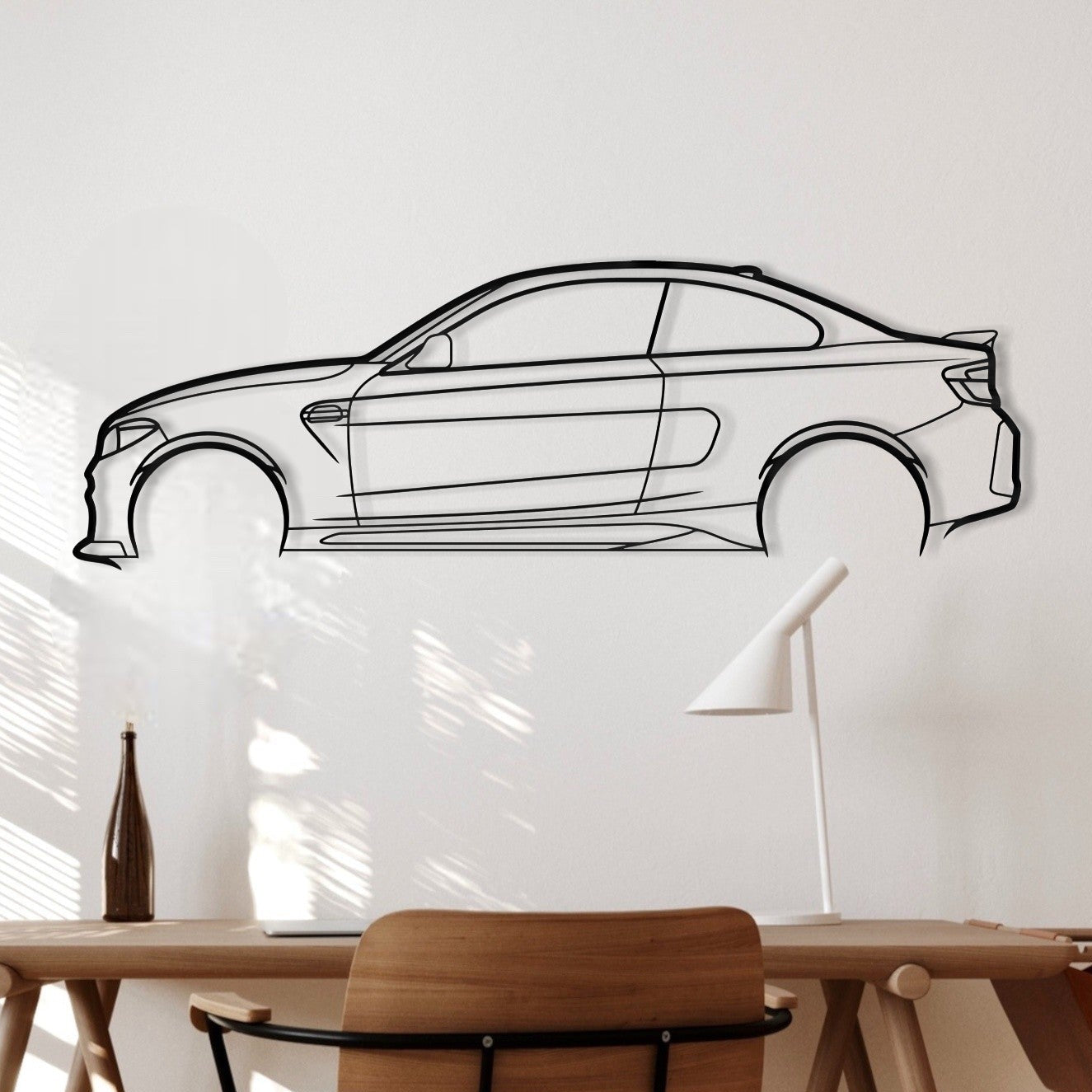 A minimalist interior scene with a BMW M2C metal wall art silhouette a white wall, wooden desk, wooden chair, and a black line-art car decal mounted above the desk.