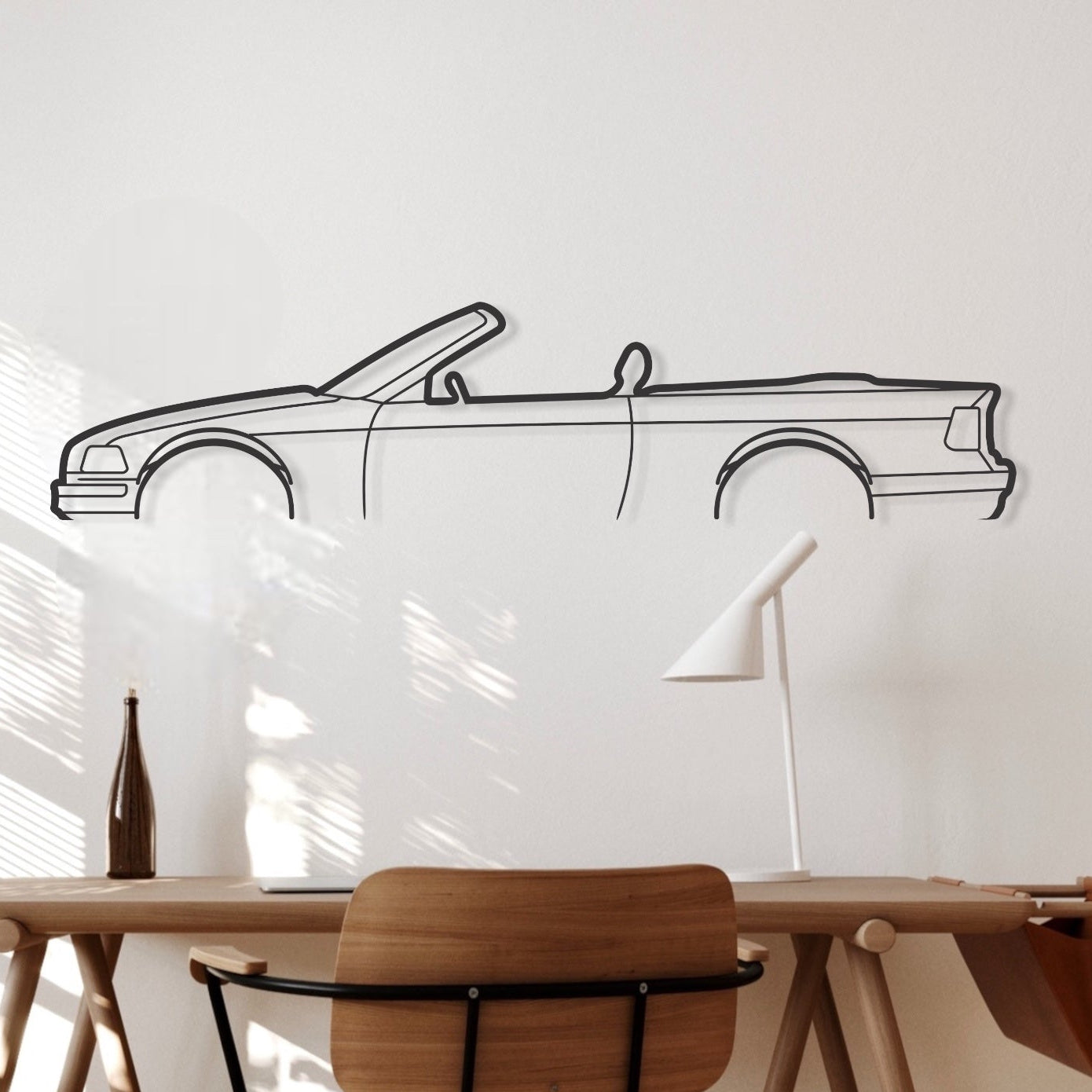 A metal silhouette of a BMW E36 Convertible car wall art, displayed on a wall above a wooden desk with a chair and lamp in a room with natural lighting.