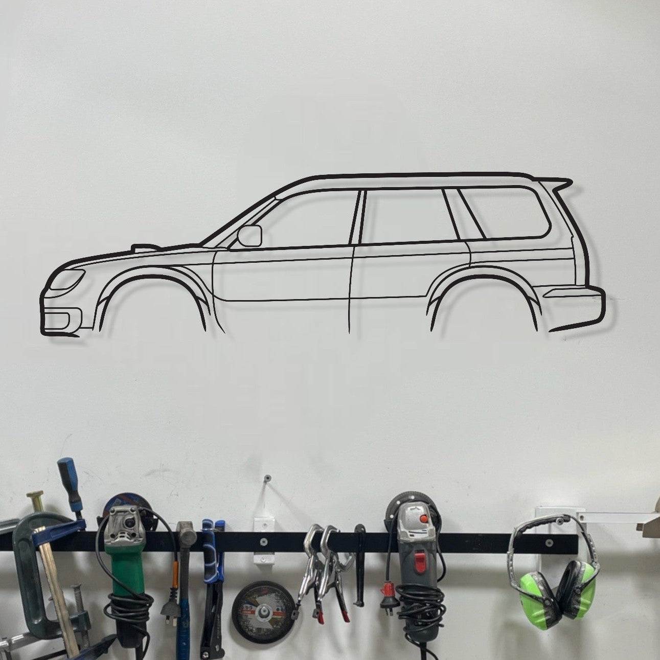 Wall-mounted tool rack with various tools and a car outline on a white wall.